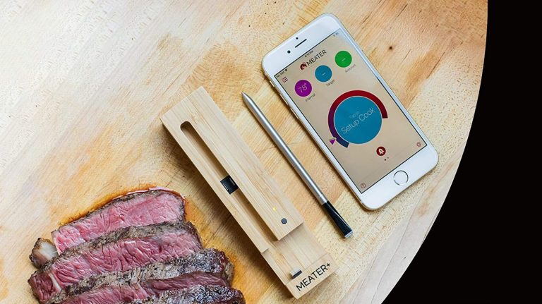 The MEATER Plus is shown next to a smartphone and some sliced steak