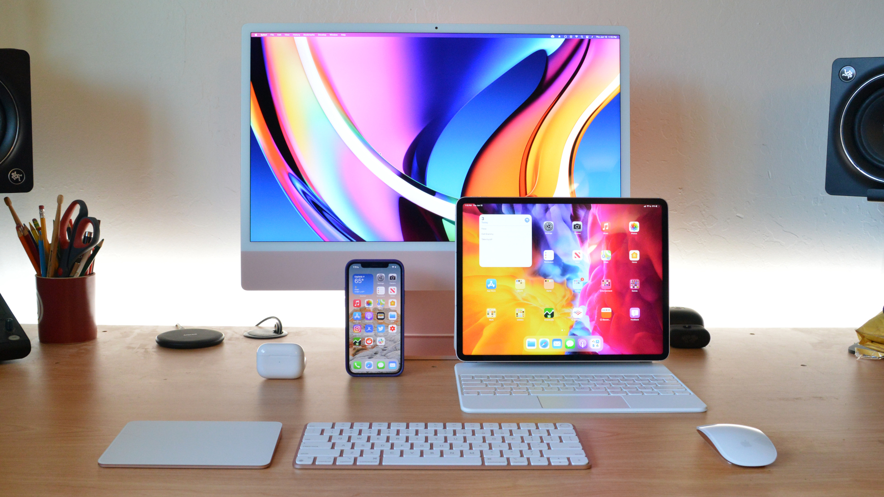 Apple iMac pictured with iPad, iPhone, AirPods, keyboard, mouse, and trackpad on a wood table.