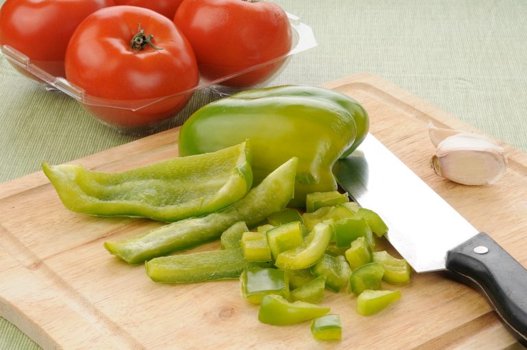 Diced green pepper on a cutting board with a container of tomatoes in the background
