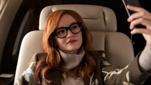 Inventing Anna young woman wearing glasses and holding up smartphone