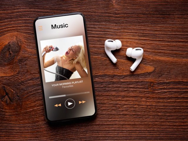 Music player on screen of iPhone and AirPods Pro on wooden surface.