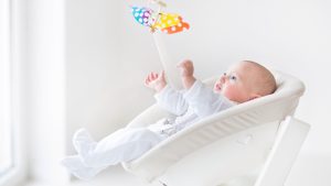 Cute newborn baby boy watching a colorful mobile toy sitting in a white chair next to a window.