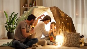 Father with torch light telling scary stories to his daughter and wife in kids tent at night at home.